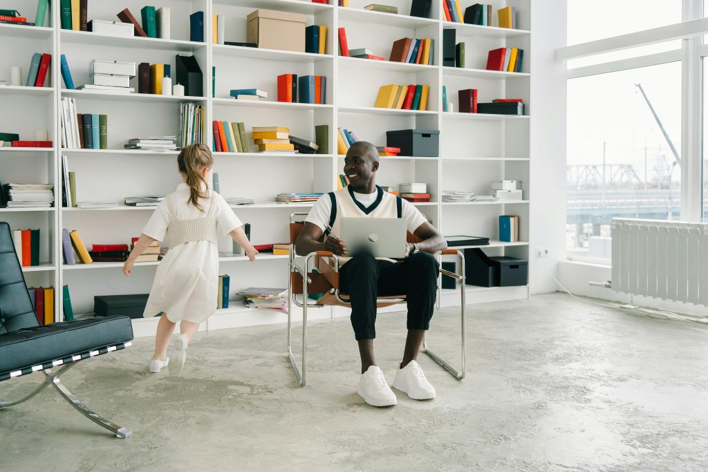 An adult sitting with a laptop in a modern library while a child plays nearby. Bright, educational setting.